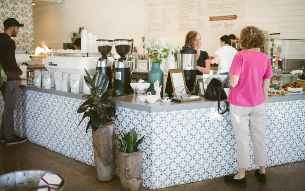 A cozy café interior with a patterned counter, coffee machines, and customers interacting. Plants add a touch of greenery.