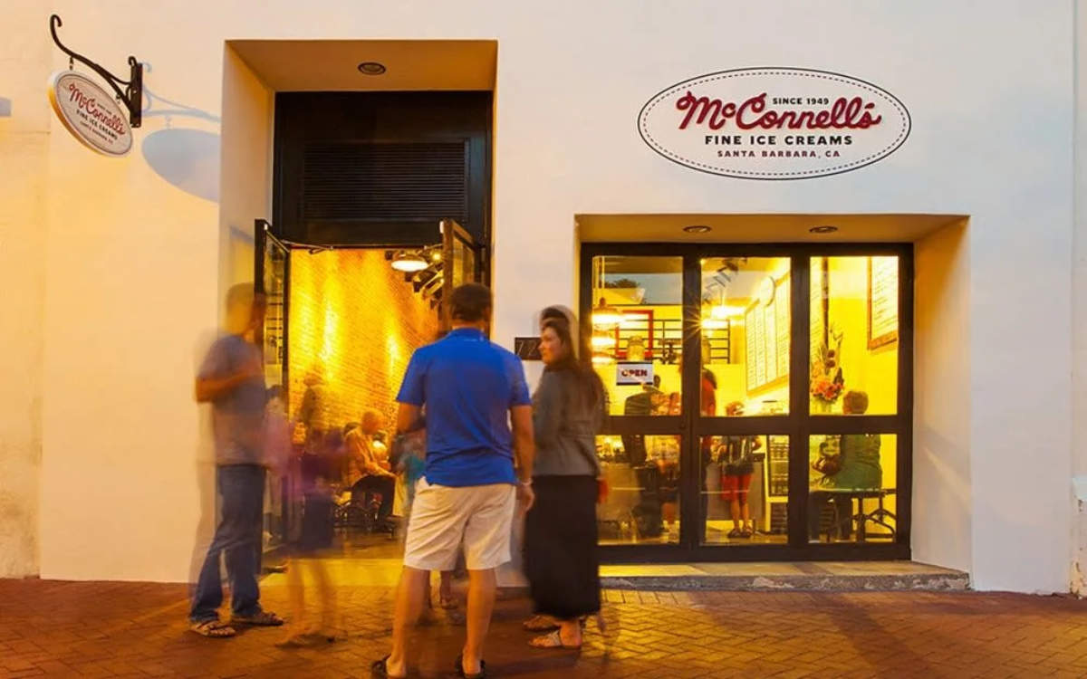 A bustling ice cream shop entrance with people outside, warm lighting visible inside, and a sign reading "McConnell's Fine Ice Creams."