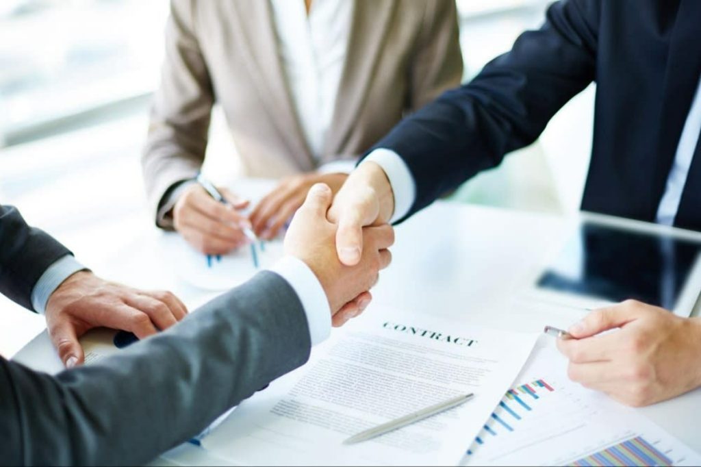 Two business professionals shaking hands over a contract on a table, with documents and a tablet in the background.