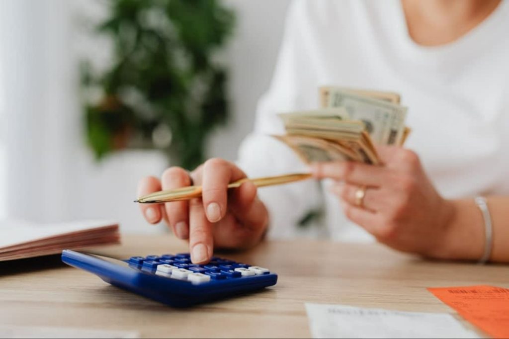 A person using a calculator while holding cash, with a notebook and receipts on a wooden table.