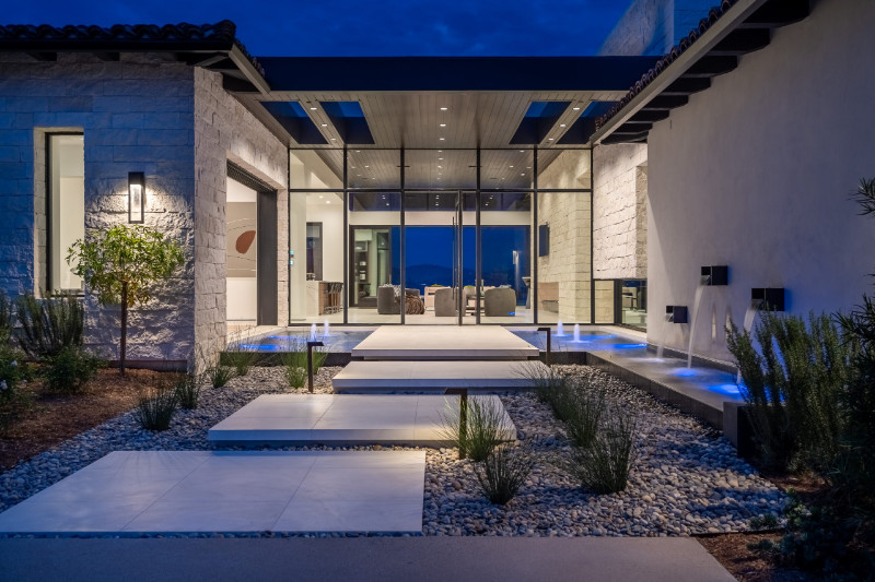 Modern luxury home exterior at night, featuring a pathway with large white stone tiles surrounded by gravel and minimal landscaping, leading to a glass entrance with sleek water features illuminated by blue lighting.