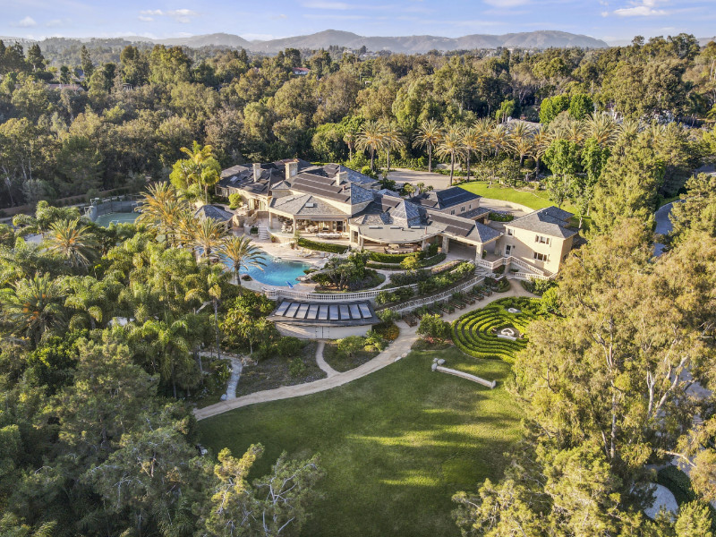  Aerial view of 16701 Camino Sierra surrounded by lush greenery, tall palm trees, and landscaped gardens. The property features a large main residence with a unique architectural design, multiple outdoor terraces, a swimming pool with a curved design, and a spacious lawn area with a decorative circular pattern.