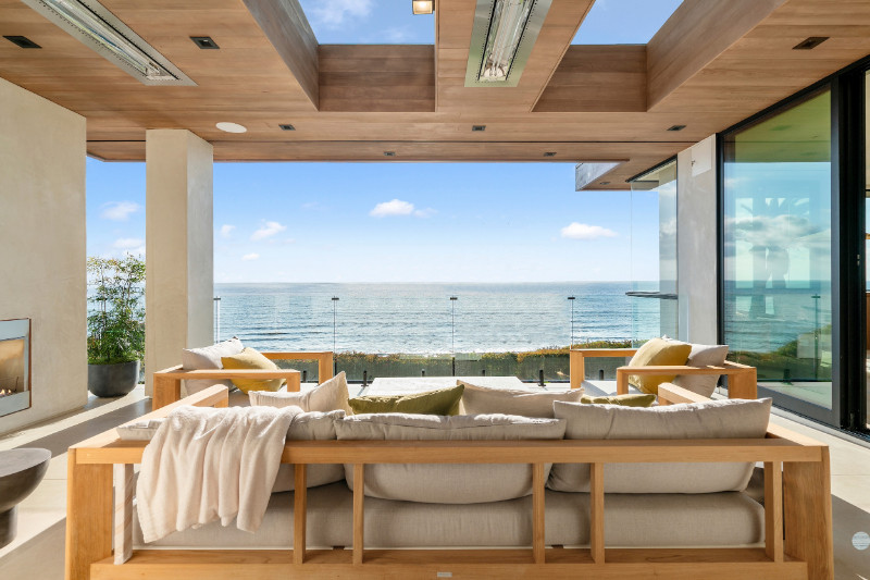 516 Stratford Ct oceanfront outdoor seating area with modern wooden furniture, soft beige cushions, and a cozy throw blanket, set under a wooden ceiling with recessed lighting.