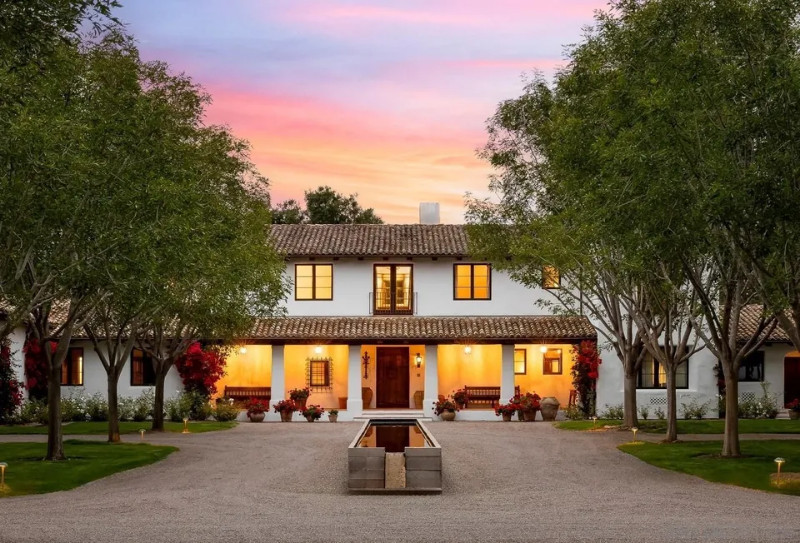 Elegant Mediterranean-style estate with a white stucco exterior, terracotta-tiled roof, and warm lighting illuminating the front porch. The home is surrounded by lush trees and colorful potted plants, with a gravel driveway leading up to the entrance.