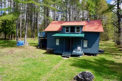 Seasonal cabin for sale in Wells NY at 958 State Hwy 30 featuring an Adirondack-style cabin with a covered front porch, metal roof, and wooded surroundings on a 0.72-acre lot.