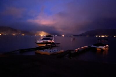 Great Sacandaga Lake Ring of Fire at dusk with shoreline bonfires glowing across the lake and boats docked in the foreground.