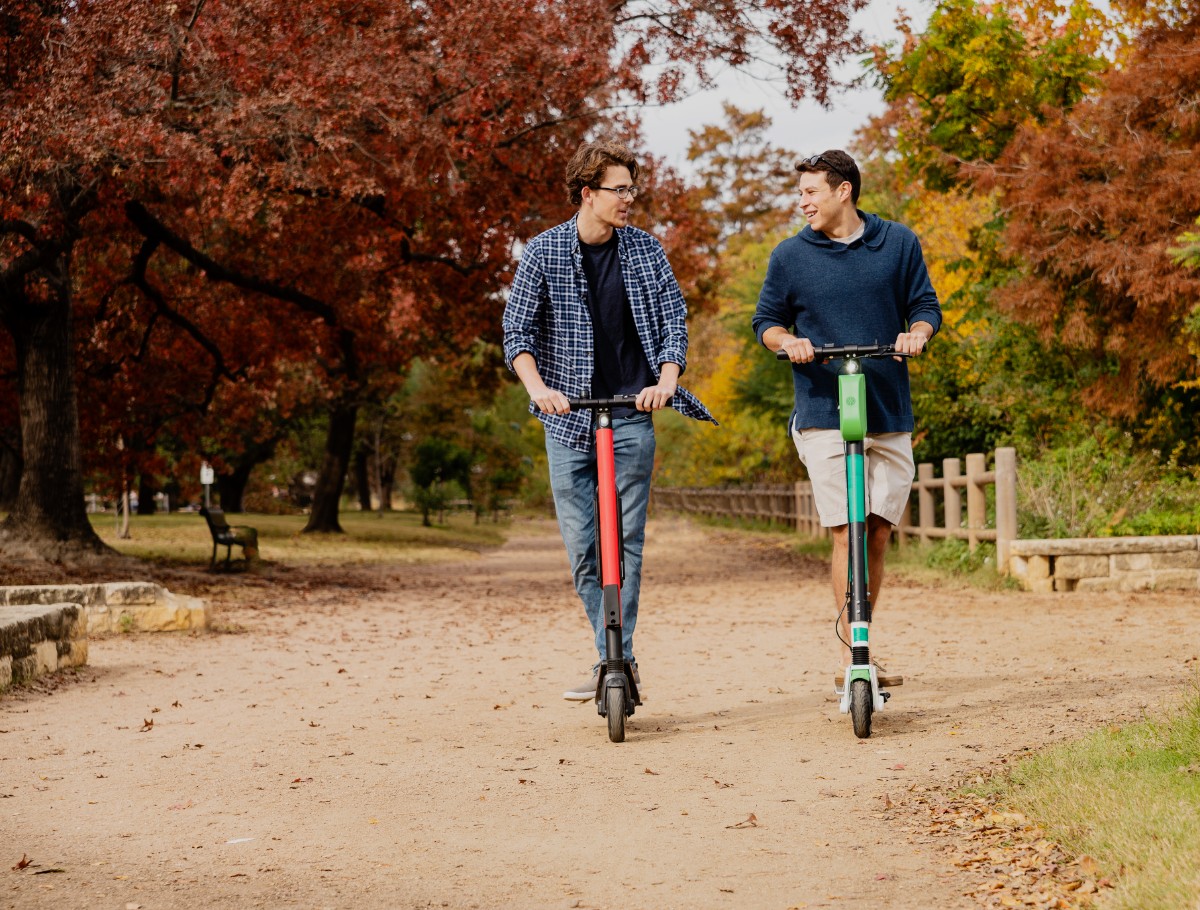 Two young, about 30-years-old, male friends, Latino, and Caucasian White, riding the rental electrical scooter in the colorful autumn park
