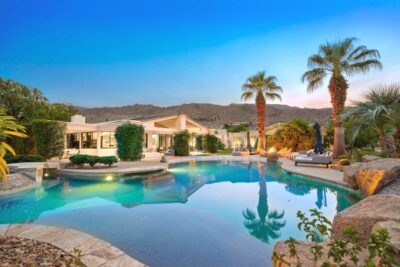 Desert modern home with a large swimming pool at sunset, framed by palm trees and mountain views