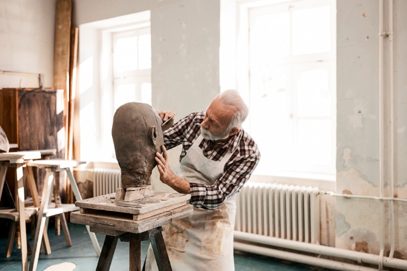 Senior man making statue of clay shaping a face with work tool