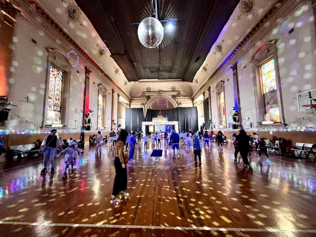 Interior View of the Roller Skating Rink at the Church of 8 Wheels, with People Skating, Disco Lights lighting up the floor and walls, and a Disco Ball Chandelier