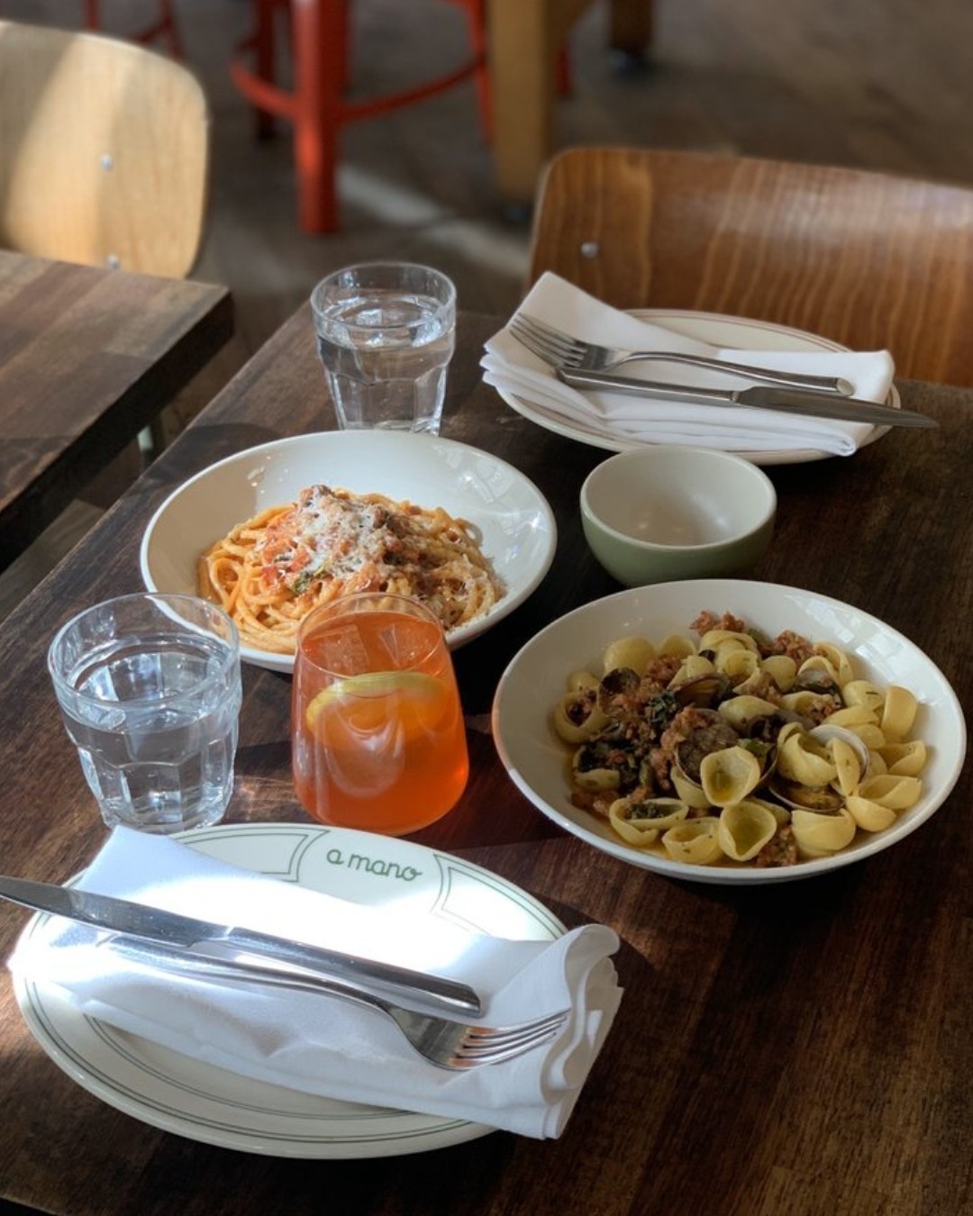 Two Plates of Pasta Dishes, an Aperol Spritz, Two Glasses of Water, and Place Settings with plate that reads a Mano