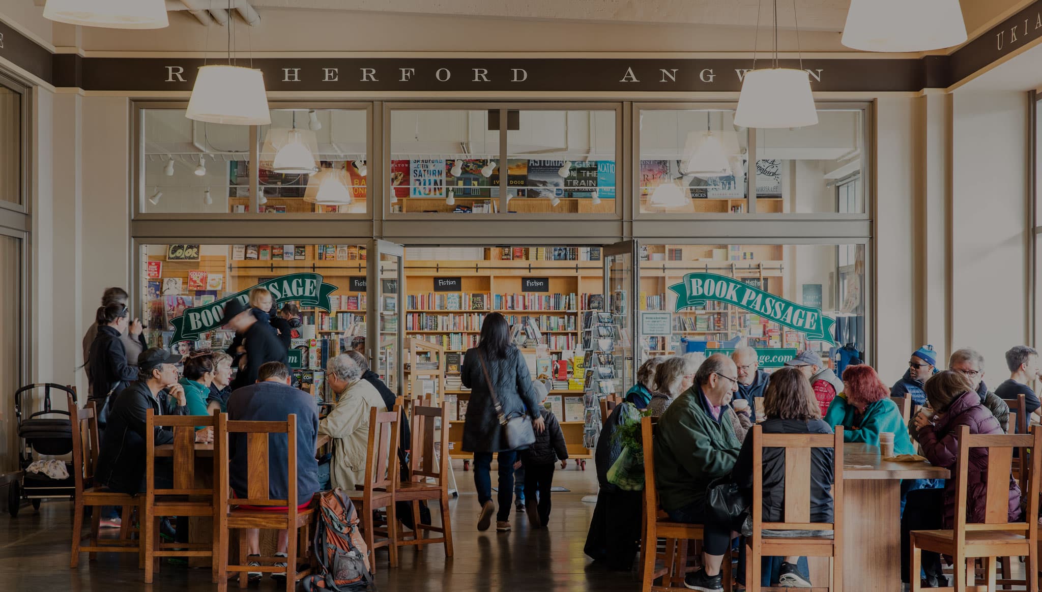 View of bustling Book Passage storefront with lots of shoppers and readers outside the store at San Francisco’s historic Ferry Building, plus lines of books against the wall in the background