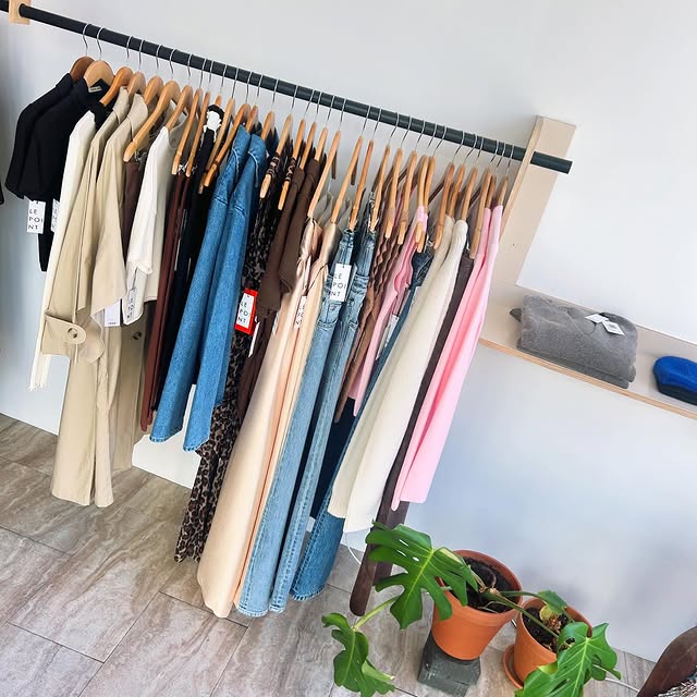 Rack of clothing items for sale at Le Point in the Mission District, including jeans, denim jackets, trench coats, black collared shirts, pink sweaters, and more