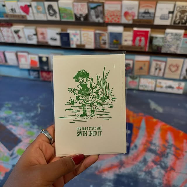 Artisan printed card with green graphic and lettering that reads “cry me a river and SWIM INTO IT”, with a large display of cards in the background