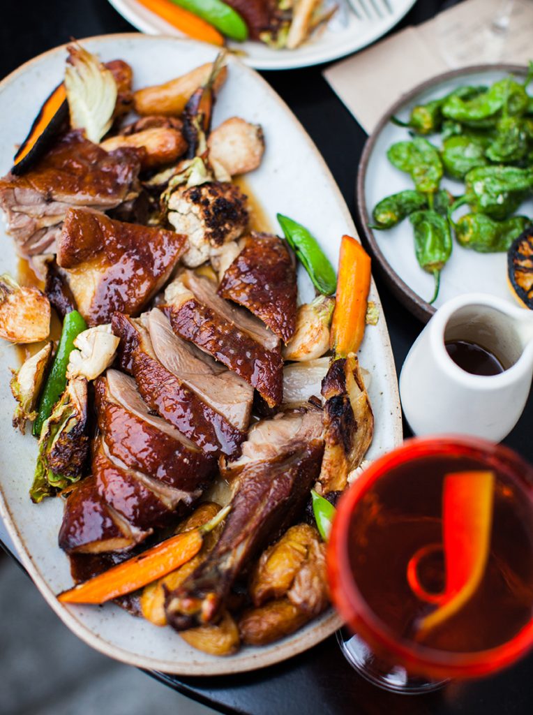 Plate of sliced smoked duck breast with roasted heirloom baby carrots, snap peas, and separate plate of shishito peppers, with a blurred Manhattan cocktail with an orange twist in the foreground