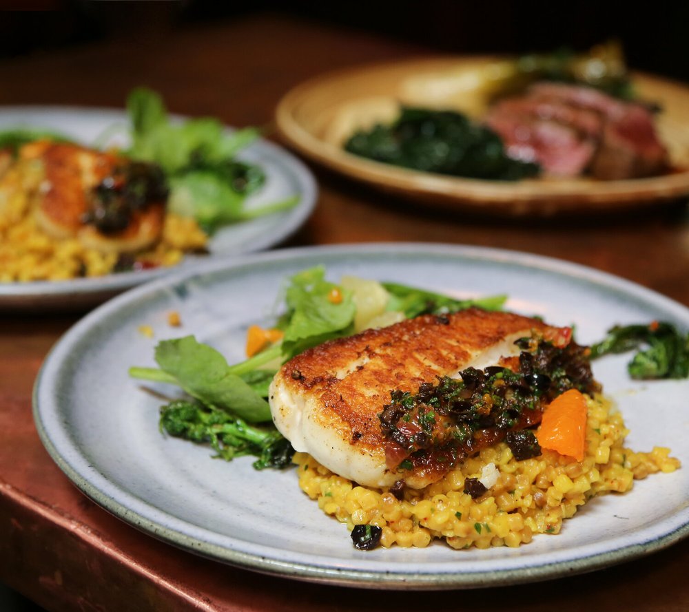 Plate of grilled pan-seared fish atop a bed of couscous, with a side of greens