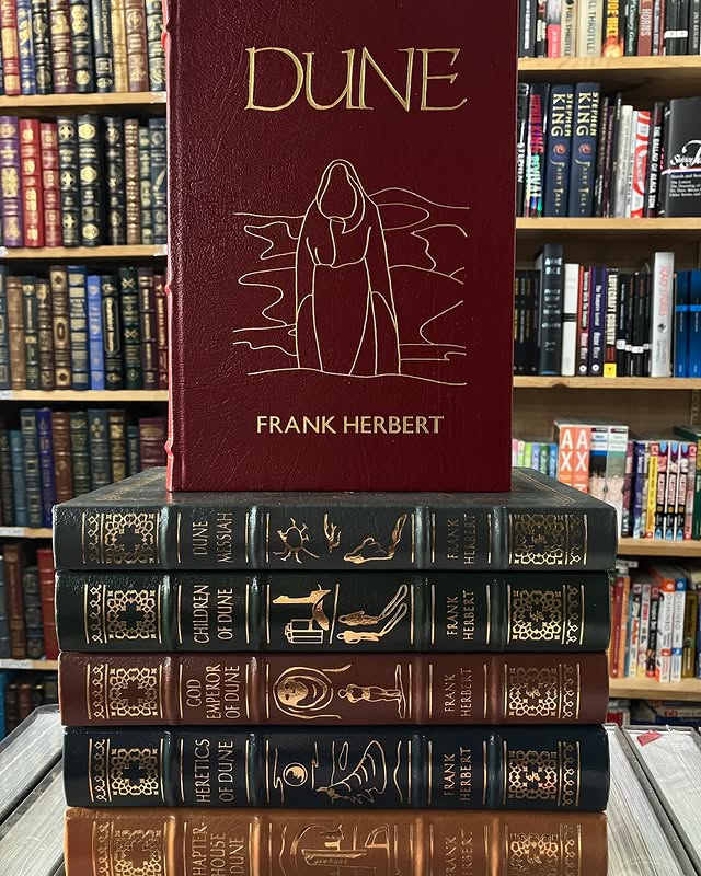 The original six volumes of the Dune series by Frank Herbert pictured in front of a row of stacked bookshelves