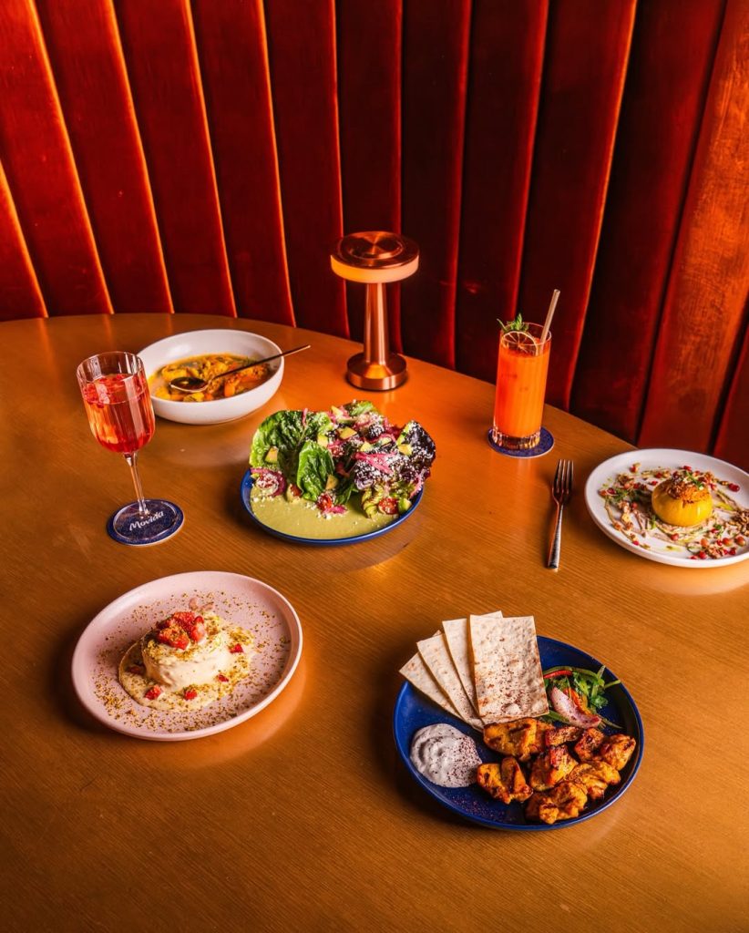 A fusion of Persian and Mexican flavors as highlighted in five different dishes including a bright salad, stuffed tomato, and flatbread with dipping sauce and grilled meat, plus two craft cocktails, set atop a light wood table with red velvet lounge booth seating