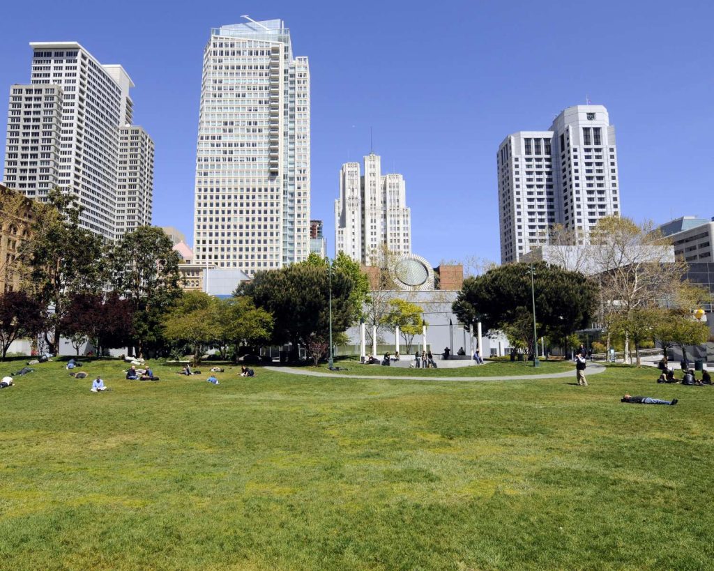 People basking in the sun on the Great Lawn at Yerba Buena Gardens, with views of SF’s skyscrapers in the background on a bright blue day