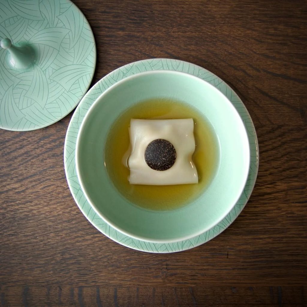 Pork and fish maw square dumpling centered in a yellow-colored ham broth on a light teal plate from Benu.