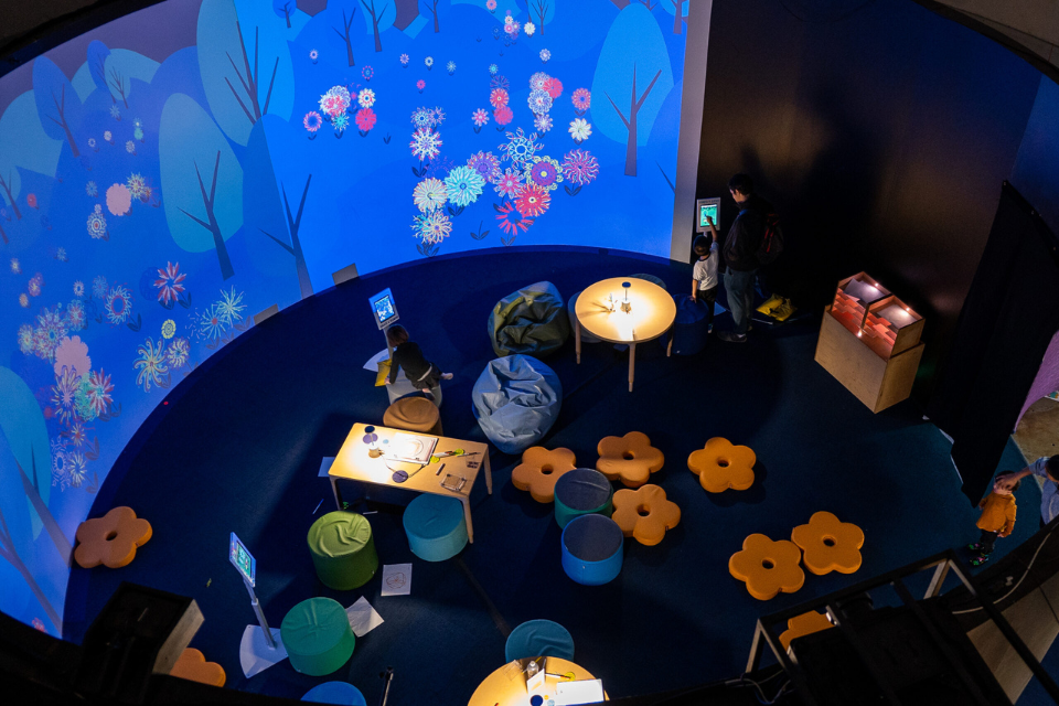 Overhead image of a dimly lit children’s play and interactive area featuring flower padded cushions, bean bags, a table, and art wall