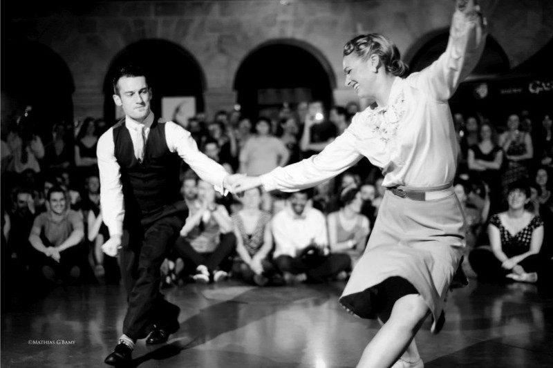 A couple performs a dynamic dance on a wooden floor, showcasing elegant movements in formal attire. Lights and decorations are in the background.