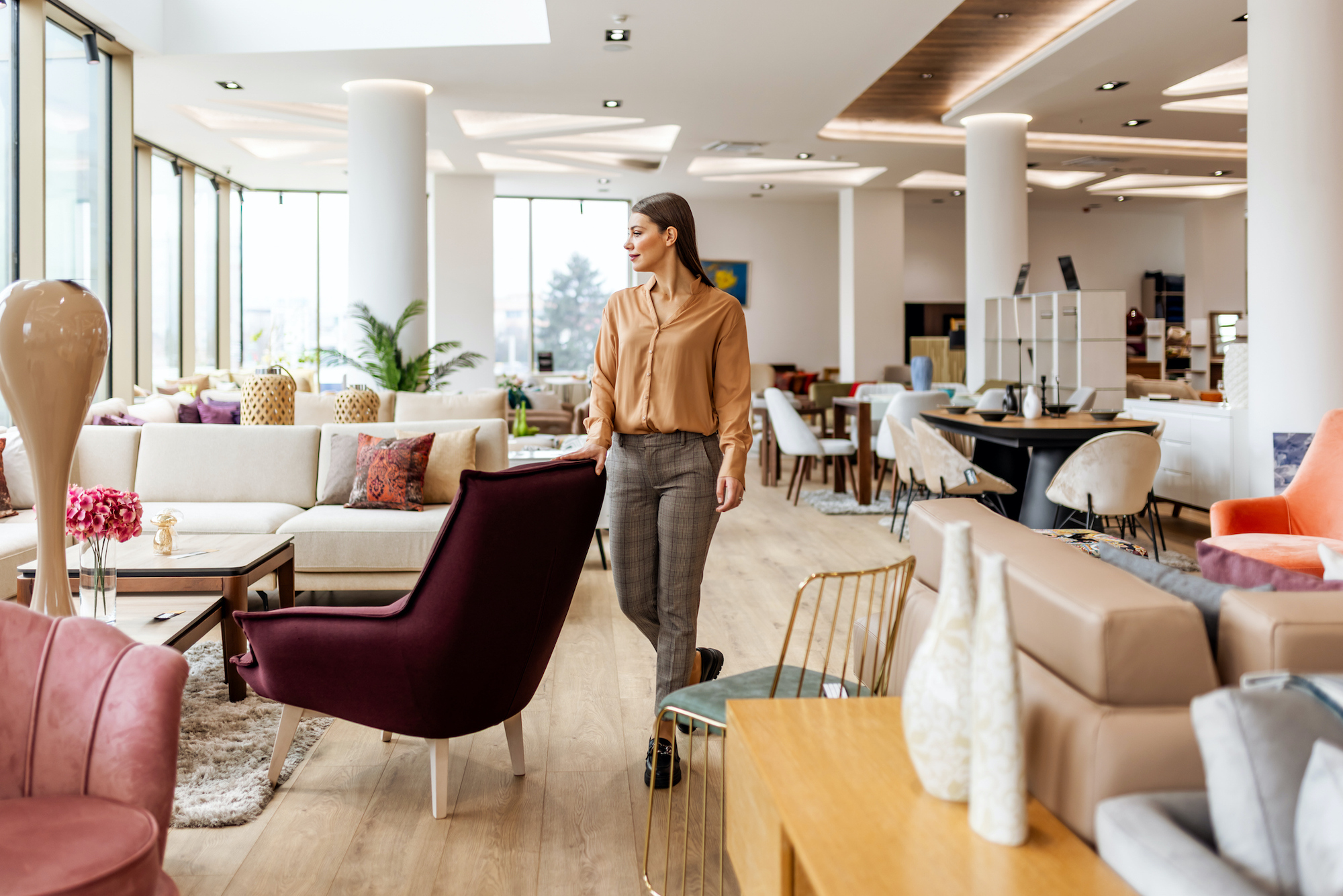 A stylish showroom full of contemporary furniture. A woman in a peach blouse and gray pants strolls past colorful sofas, chairs, and accessories, highlighting interior design, home decor, and shopping vibes in a bright, airy space.