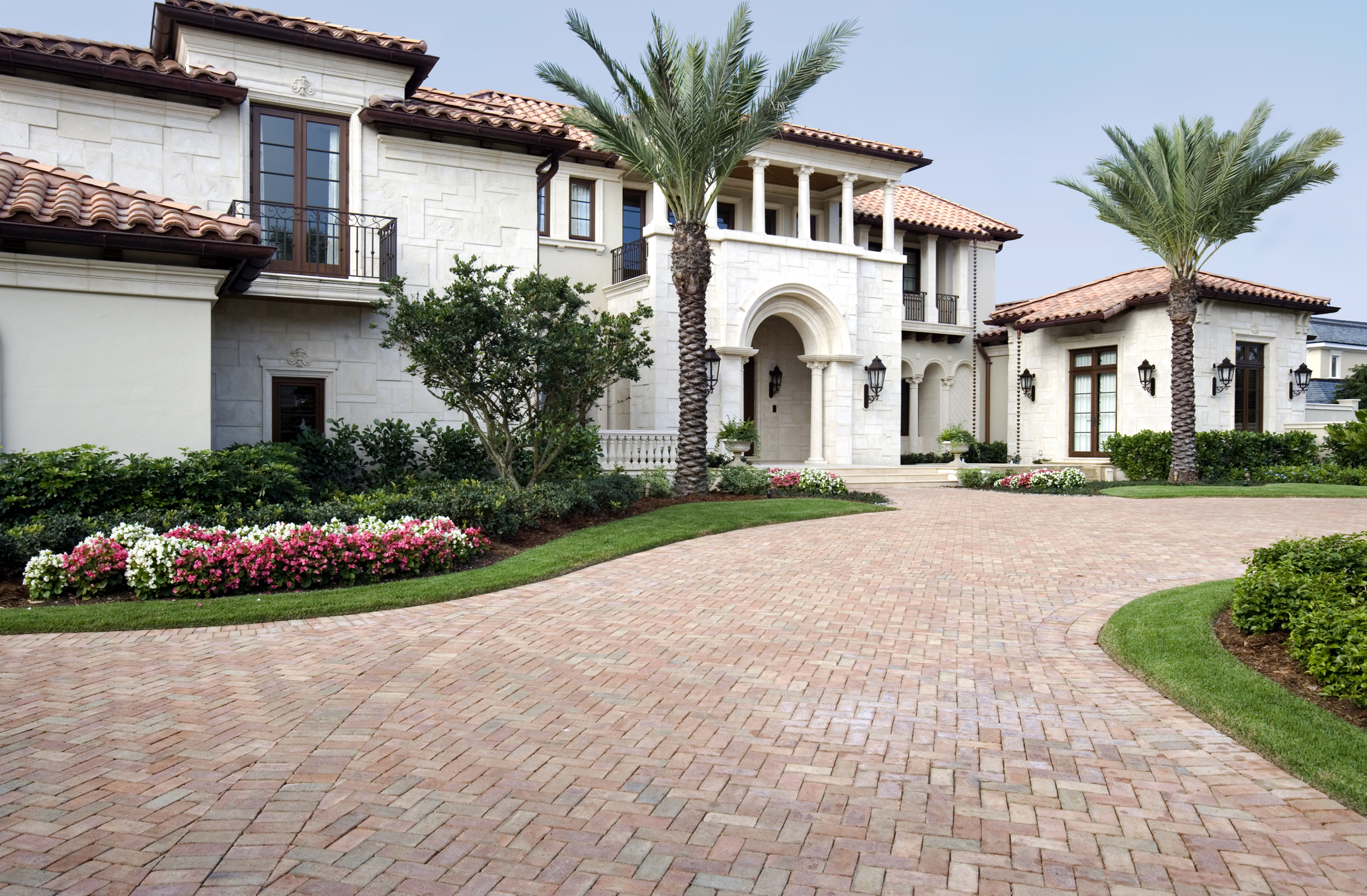 A driveway leads to a Mediterranean-style white house, a blue sky in the background and landscaping in the foreground.
