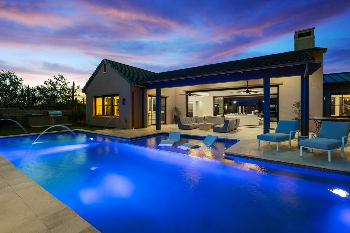 Evening view of an illuminated outdoor pool in White Horse, with lounge chairs, a covered patio area, and seating, set against a twilight sky.