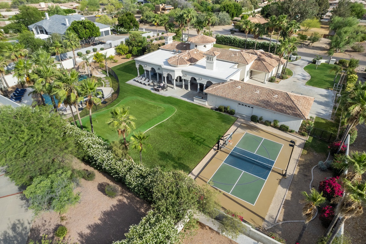 Aerial view of a luxury estate featuring a backyard with a putting green, tennis court, pool with a waterslide, and lush landscaping surrounded by palm trees.