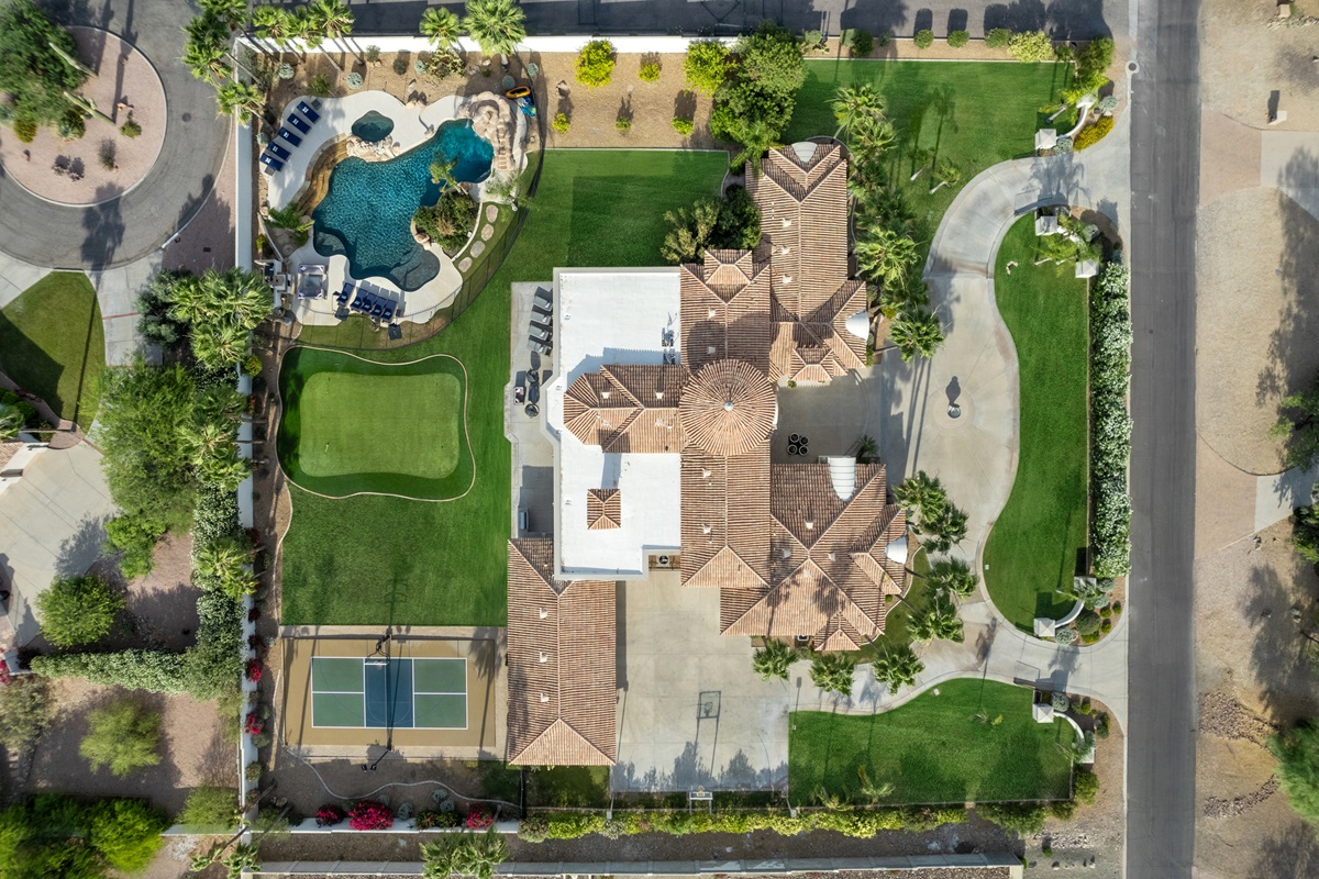 Aerial view of a large luxury estate with a pool, spa, putting green, and tennis court, surrounded by landscaped greenery and palm trees.