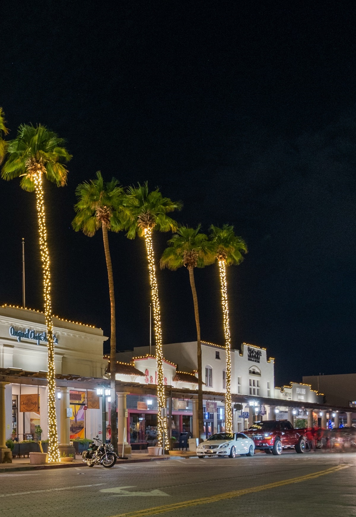 Palm trees wrapped in lights line a lively downtown street in Chandler, Arizona, with brightly lit storefronts and parked cars at night.