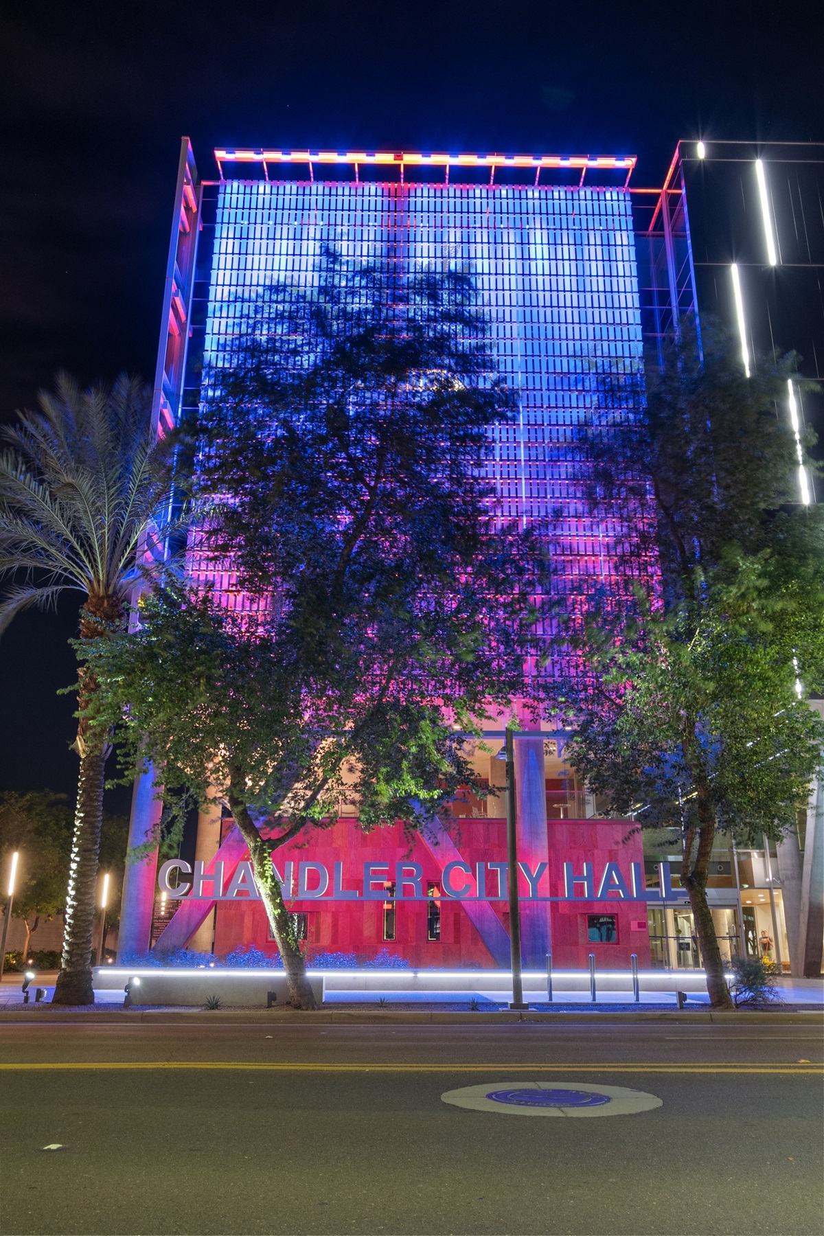 The Chandler City Hall building illuminated with colorful blue and red lights, surrounded by trees and palm trees, creating a vibrant night scene.
