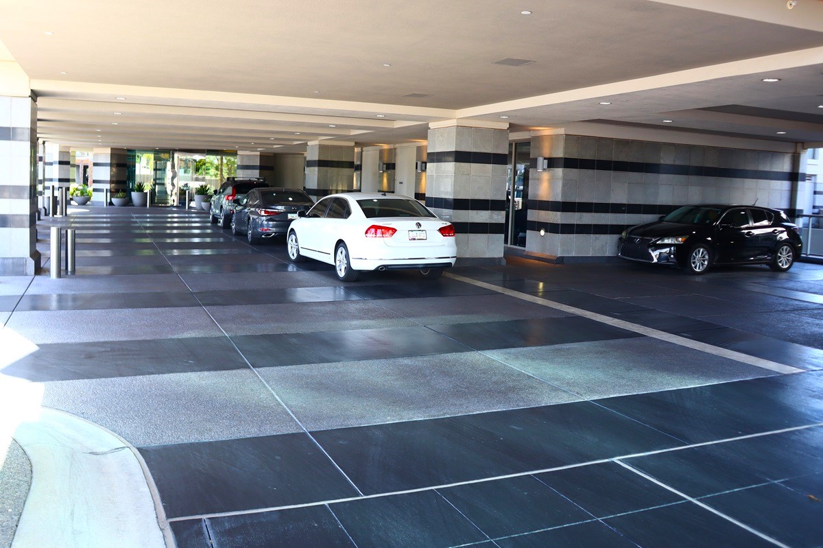 Covered driveway and entrance area at Esplanade Place, with cars parked along the side near large stone columns and potted plants.