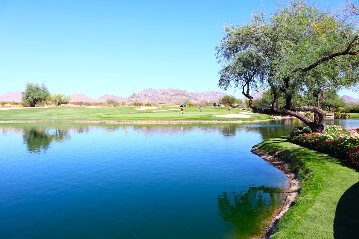 Scenic golf course with a pond, desert mountains, and a few golfers playing under a bright blue sky. 
