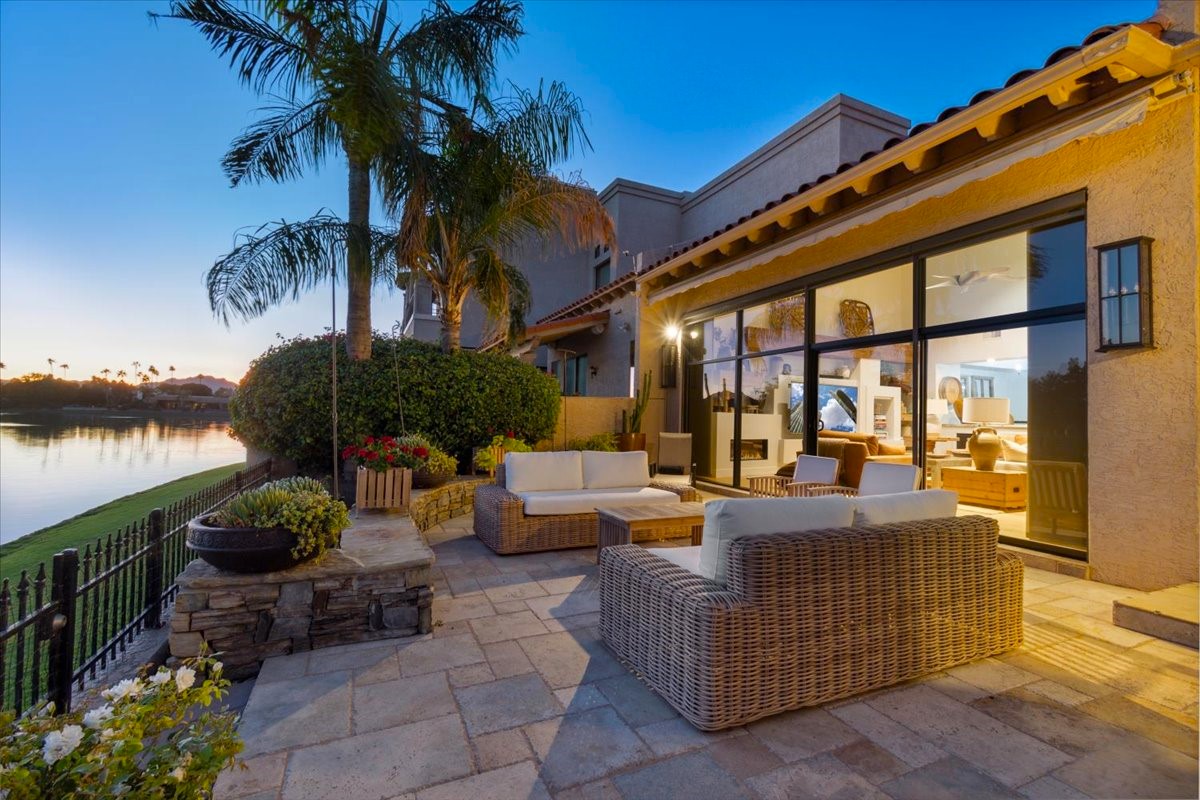 A cozy outdoor patio with wicker furniture overlooks a calm lake at sunset, framed by lush palm trees and soft outdoor lighting for a relaxing ambiance.