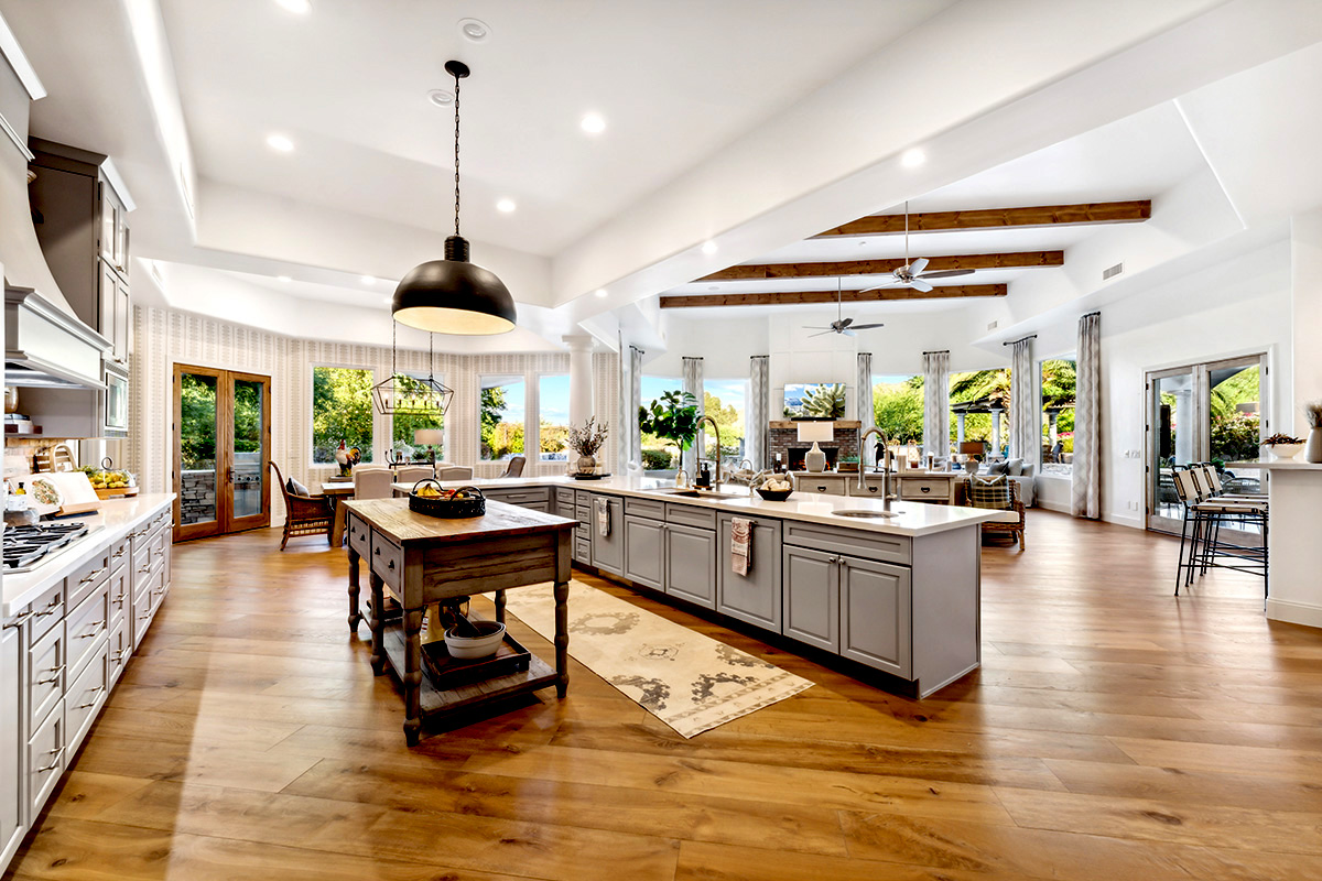 A bright, open kitchen with an oversized island, stainless steel appliances, exposed wooden beams, and large windows offering views of a landscaped backyard.
