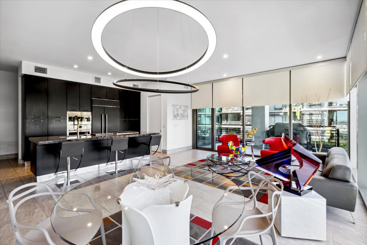 Modern luxury condo living area featuring sleek black kitchen cabinets, open concept dining with glass table, bold red accent chairs, and a contemporary glass piano near floor-to-ceiling windows overlooking a private balcony.