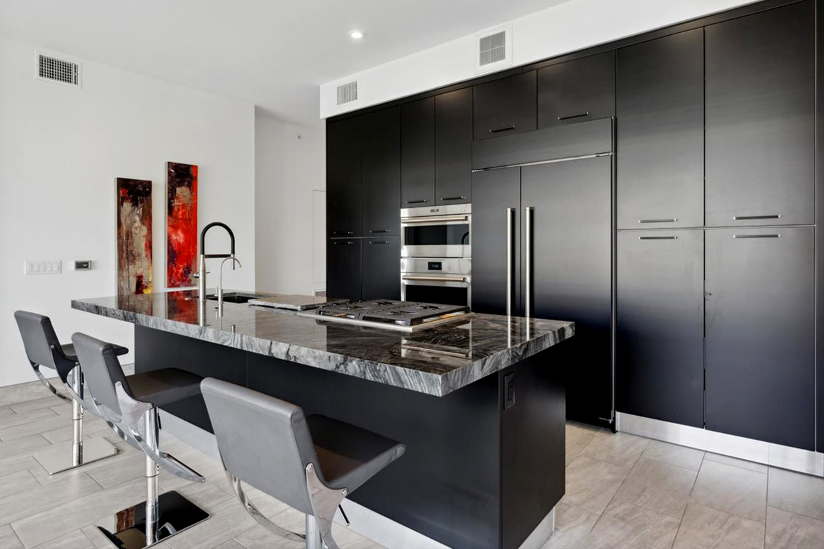 Sleek modern kitchen with black cabinetry, a large marble island with built-in gas cooktop, stainless steel appliances, and three chrome barstools, accented by abstract red artwork on the wall.