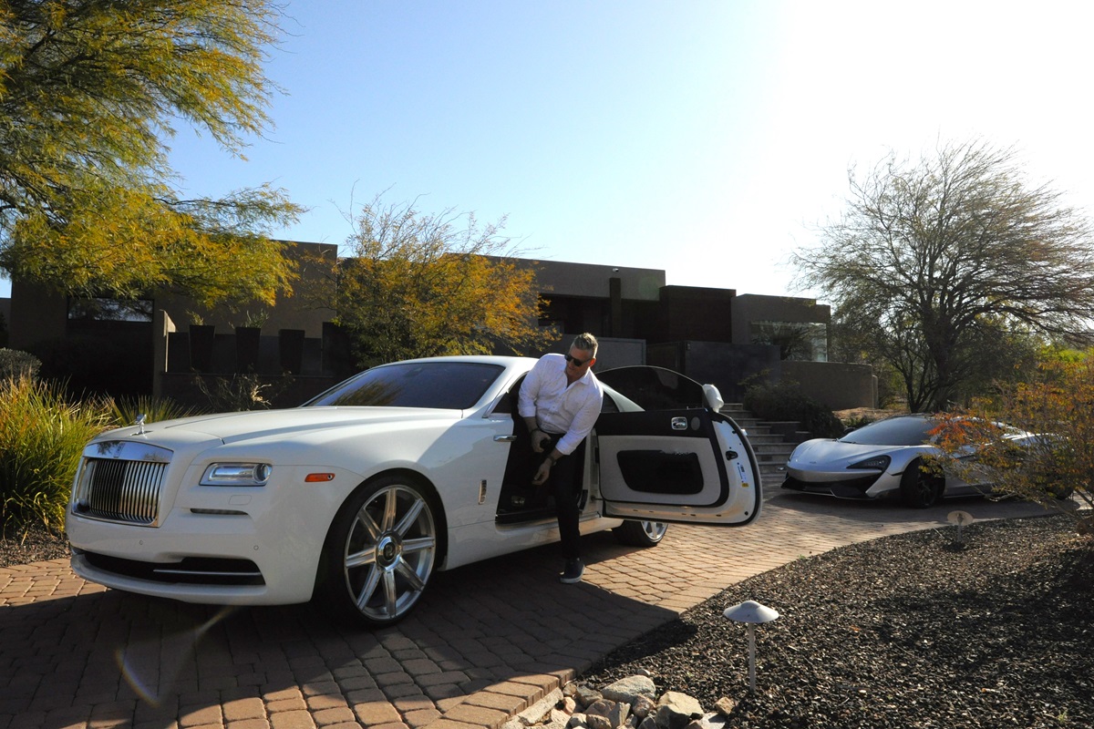 A man steps out of a white Rolls-Royce parked on a driveway, with a sports car visible in the background.