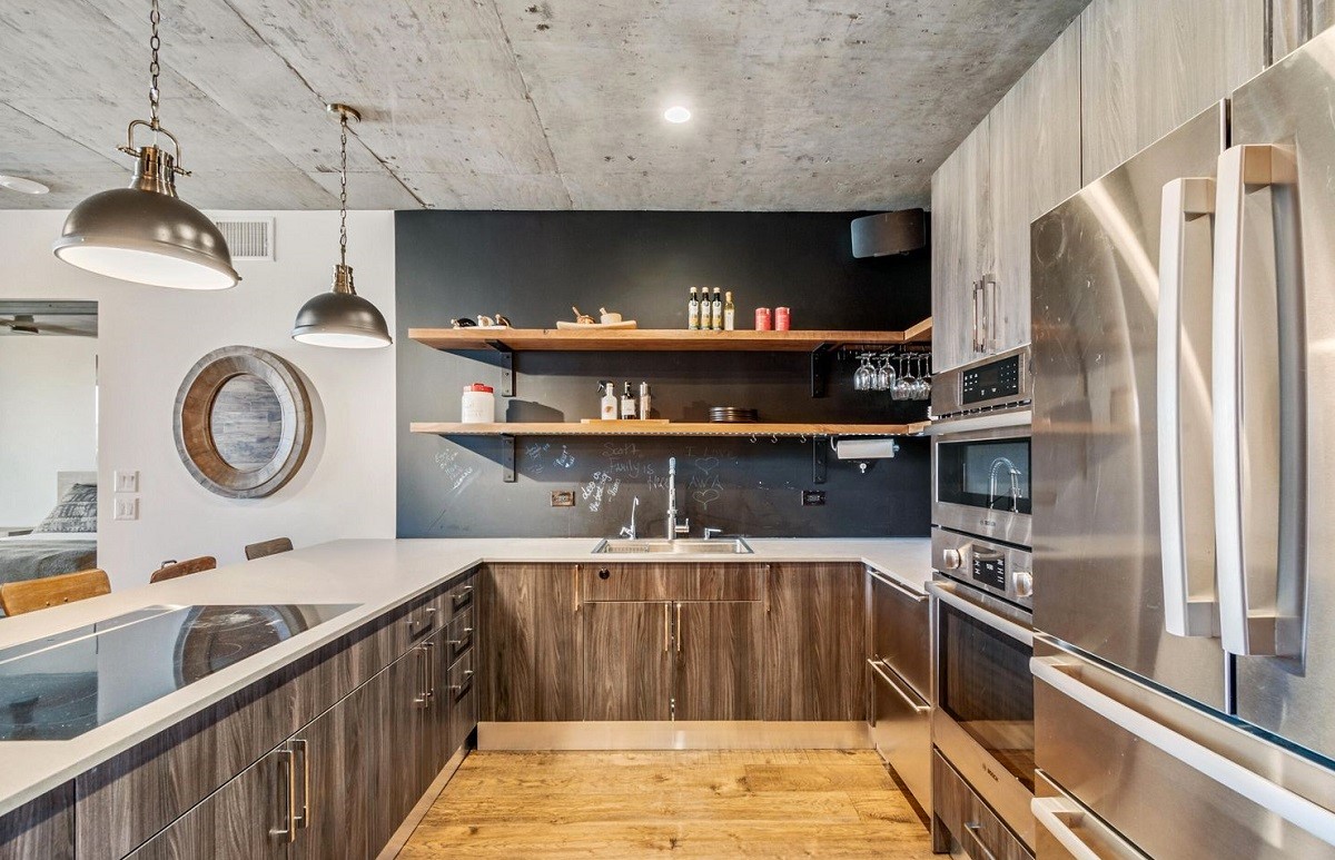 Modern industrial-style kitchen at Optima Biltmore, featuring open shelving, stainless steel appliances, pendant lighting, and wood cabinetry under a concrete ceiling.
