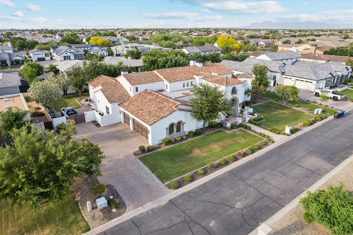 Aerial view of a large luxury home with a Spanish-style tile roof, white exterior, and landscaped front yard in a suburban neighborhood with tree-lined streets.