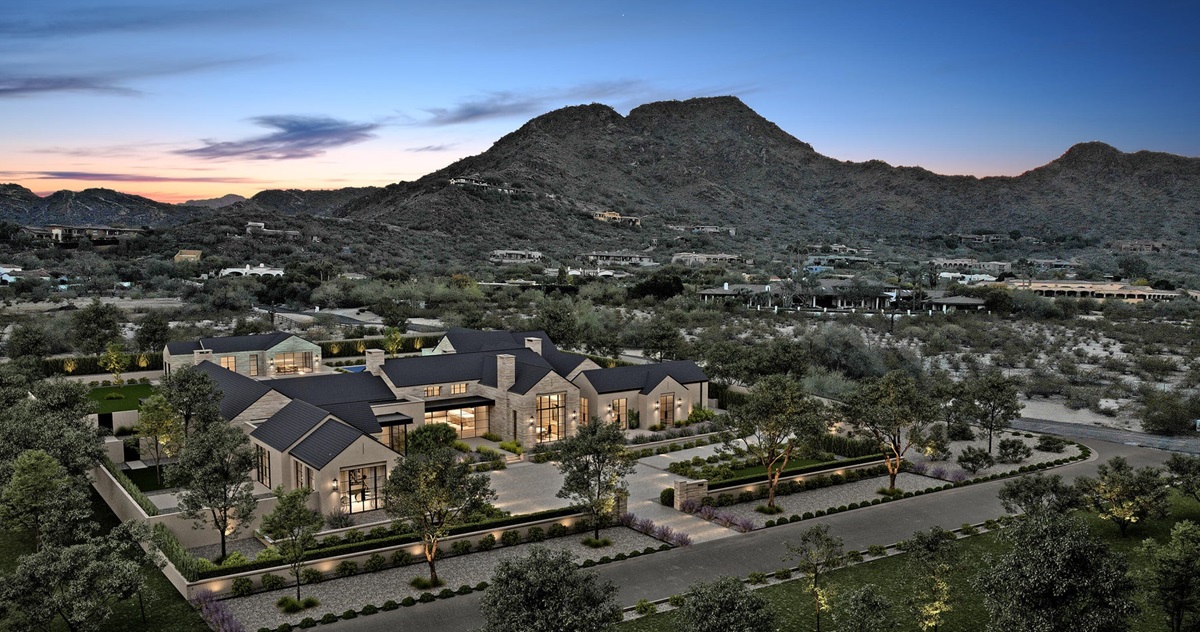 A large, elegant estate with a traditional architectural style, surrounded by greenery, nestled at the base of desert mountains, captured during twilight.
