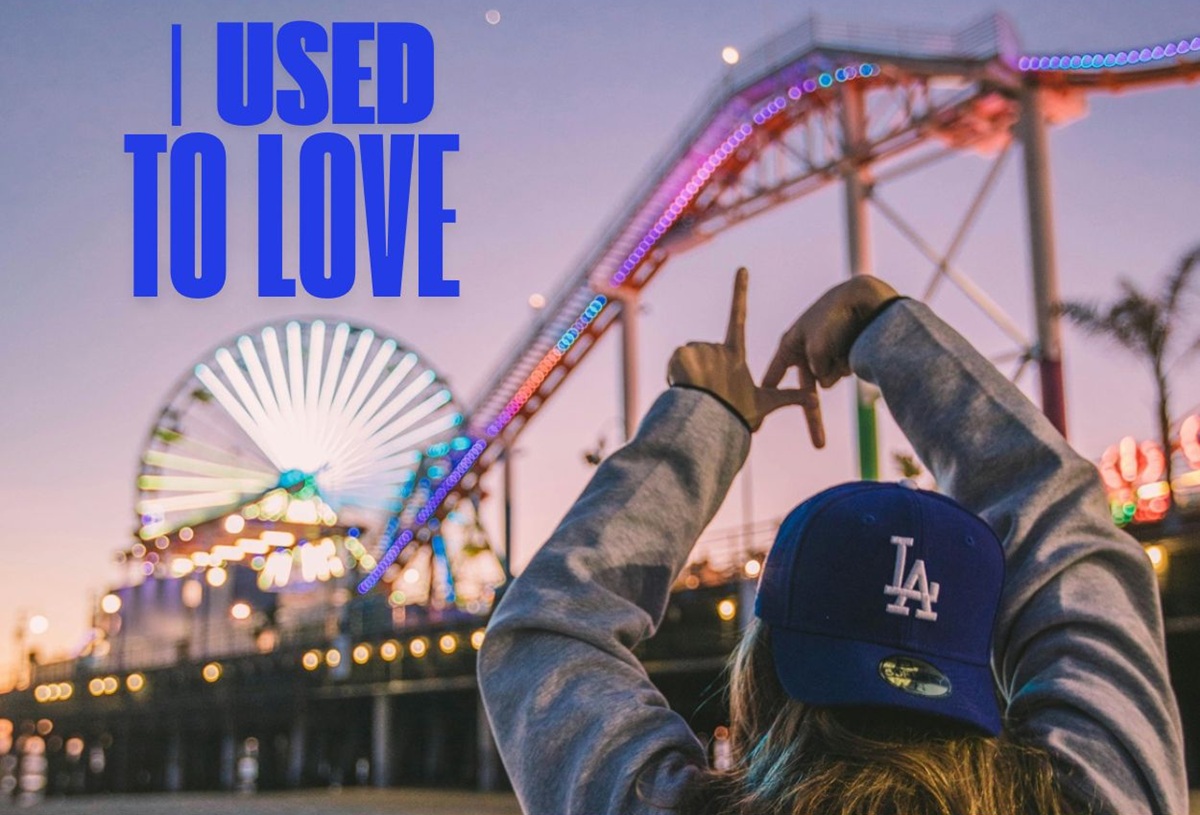 A person wearing an LA cap making a peace sign in front of a lit-up Santa Monica Pier at sunset.