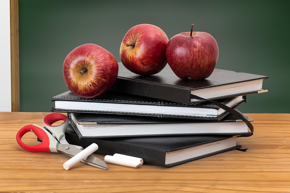 Three red apples placed on top of a stack of books, along with chalk and scissors, in front of a chalkboard.