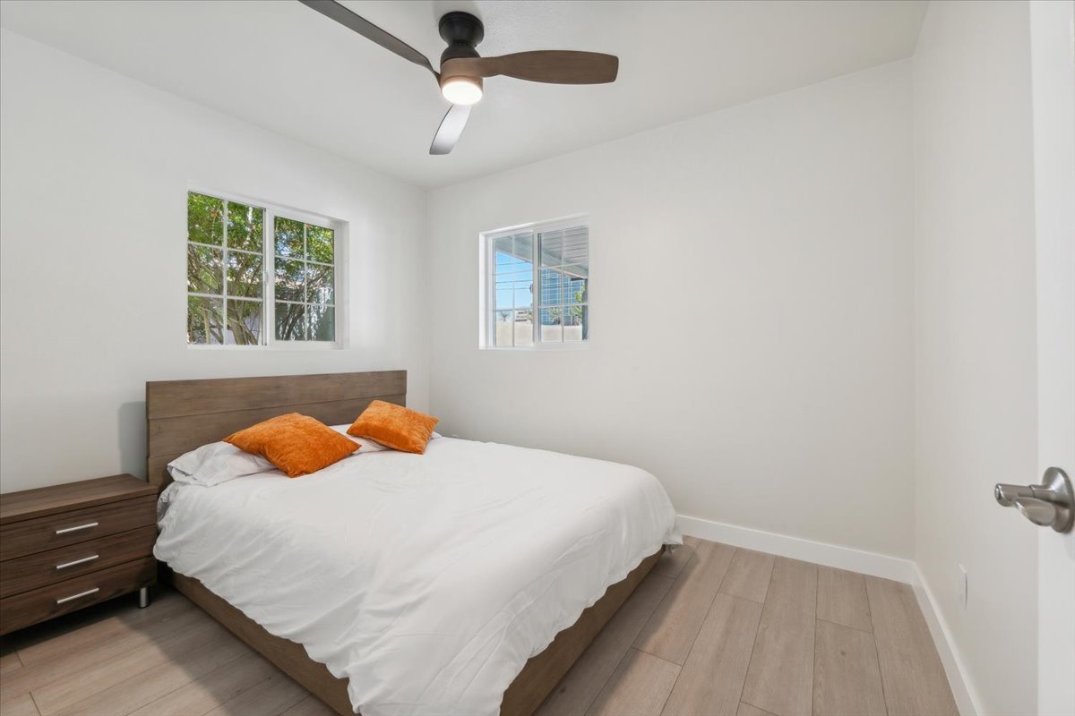 Minimalist room with a bed, orange pillows, wood headboard, side table, and ceiling fan. Bright windows.