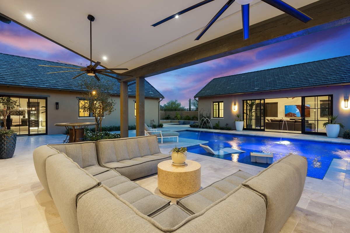 Outdoor lounge area with modern seating, ceiling fans, and a view of a pool and guest house at sunset.