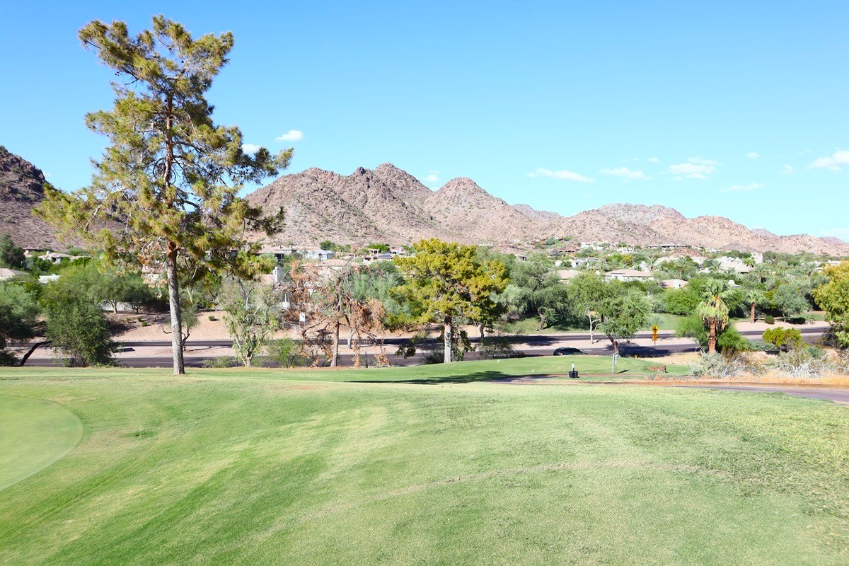 Golf course with mountain views, trees, and nearby residential homes under a bright blue sky.