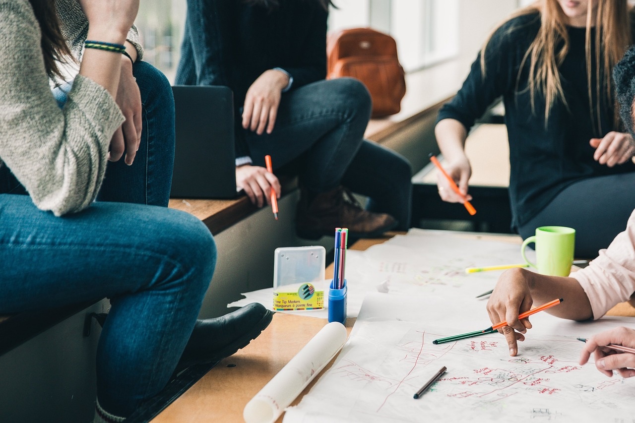 A group of students casually sitting and discussing while working on a shared project with scattered paper and colorful pens.