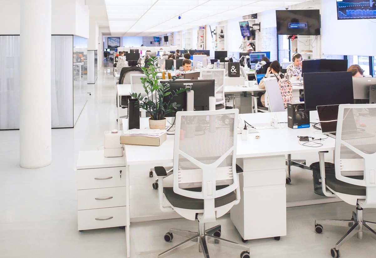 A bright, open-plan office with rows of white desks, modern chairs, computer monitors, and plants, creating a collaborative workspace.
